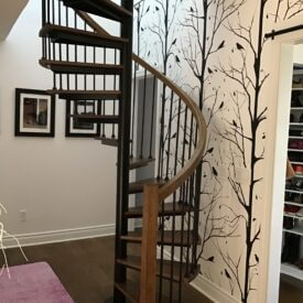 Spiral staircase with wood and black iron parts, in front of a wall with a black tree mural